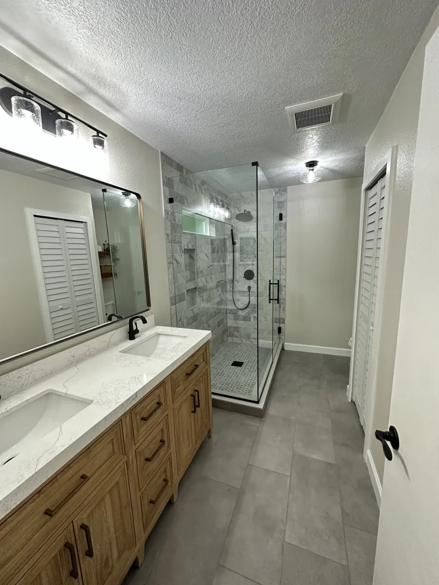 Finished bathroom with a wood double vanity, marble-look tile shower, and glass enclosure